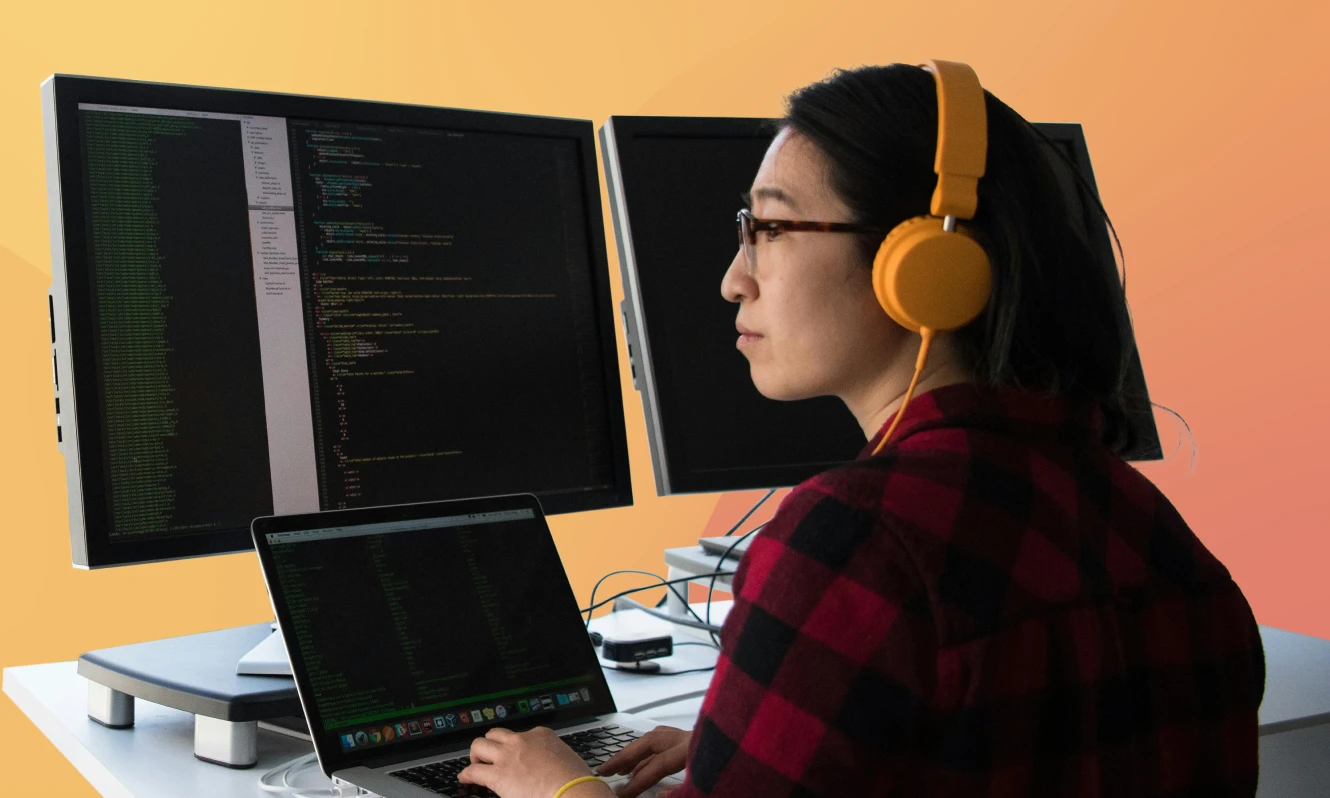 Entwicklerin mit Kopfhörern programmiert an einem Laptop mit zwei Monitoren.